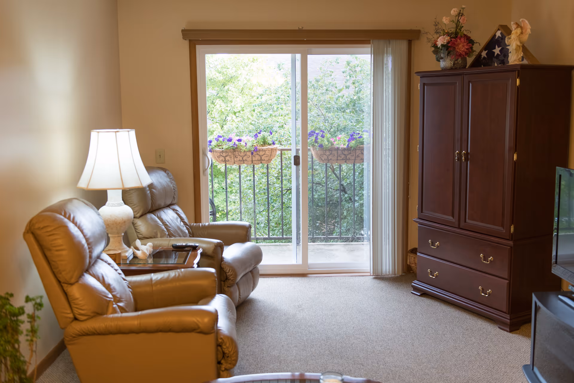 A cozy living room with two beige leather recliners, a side table with a lamp and a remote control, a wooden cabinet with decorative items on top, and a sliding glass door leading to a balcony with flower boxes and green trees outside.