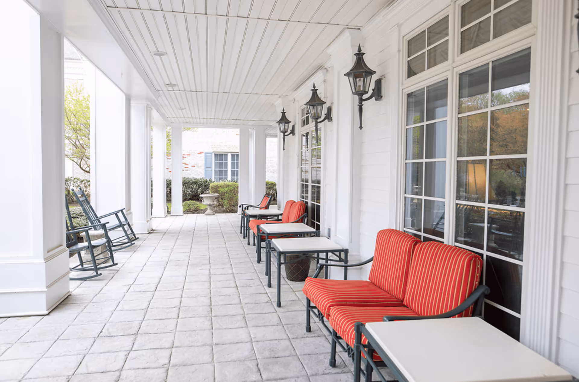 Covered outdoor patio area with white tiled floor and white ceiling. There are several black metal chairs with red striped cushions and small white tables arranged along the right side near large windows. On the left side, there are black rocking chairs. The patio is supported by white columns and has black wall-mounted lantern-style lights.
