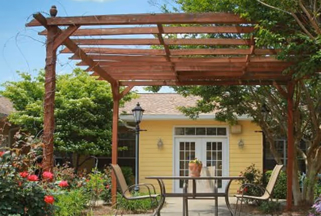 Outdoor patio area with a wooden pergola, metal table and chairs, surrounded by green trees and flowering plants in front of a yellow building with white-framed double doors.