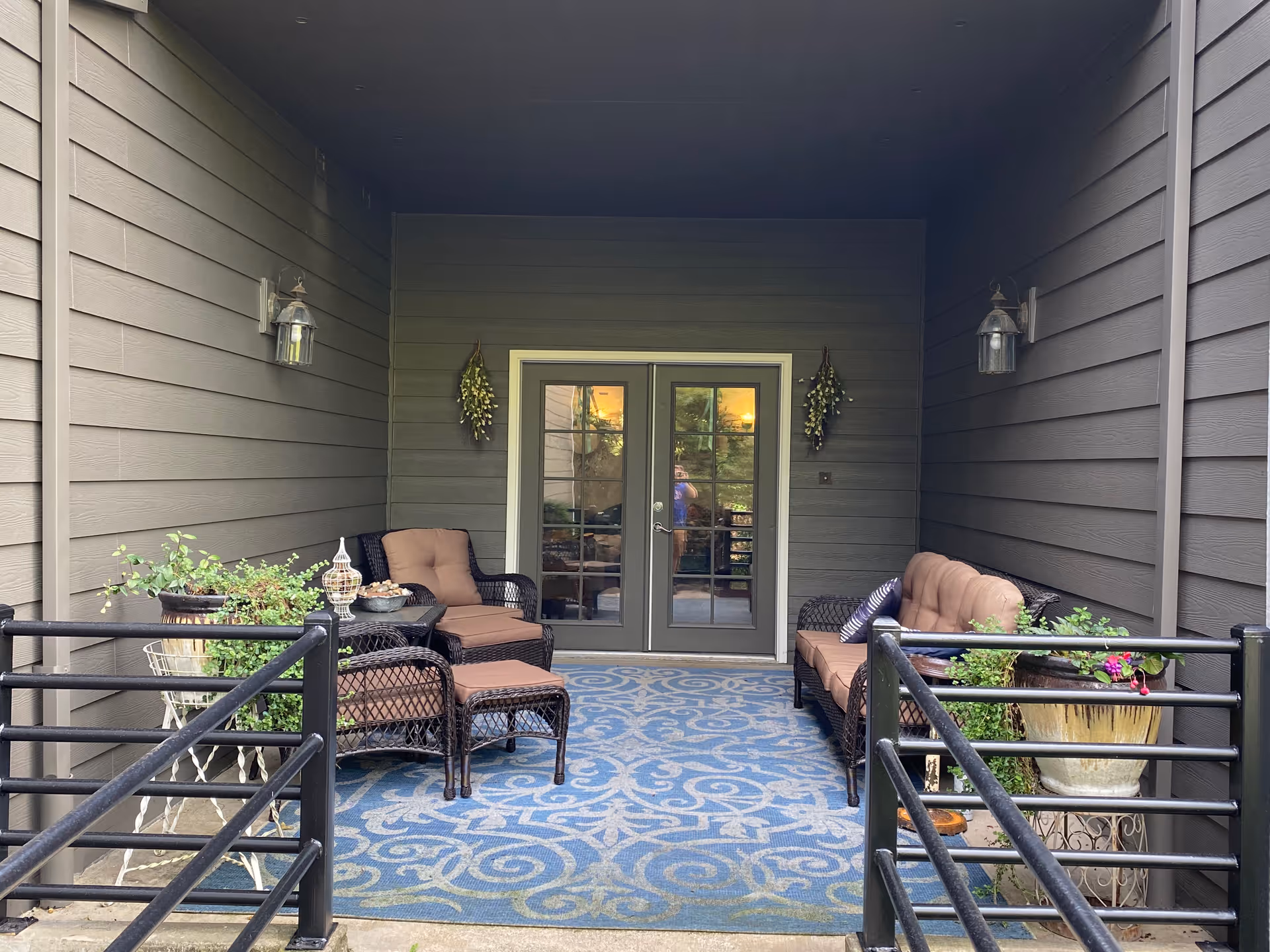 Covered outdoor patio area with cushioned wicker seating including a chair with ottoman and a sofa, a blue patterned rug, potted plants, and double glass doors leading inside. The walls are gray with two wall-mounted lantern-style lights and decorative hanging plants on either side of the doors.