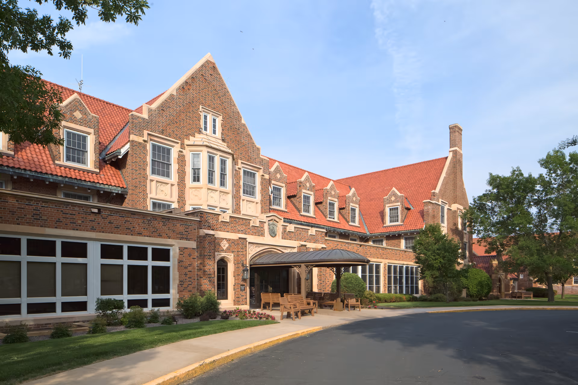 Exterior view of a large brick building with a red tiled roof, multiple windows, and a covered entrance with benches outside. The building is surrounded by trees and greenery under a clear blue sky.