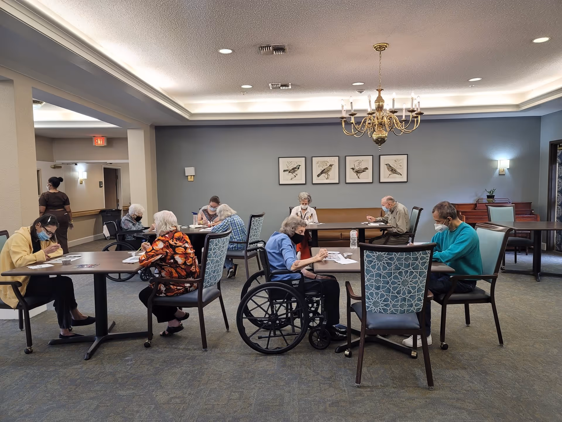 A group of elderly people sitting at tables in a well-lit room, engaged in activities such as writing or drawing. Some individuals are wearing masks, and one person is in a wheelchair. The room has a chandelier, framed bird artwork on the wall, and a piano in the background.