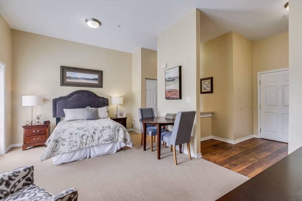 A cozy senior living bedroom featuring a bed with a dark upholstered headboard, floral bedding, and two bedside tables with lamps. Adjacent to the bed is a small dining area with a round wooden table and two gray upholstered chairs. The room has beige walls, carpeted flooring in the bedroom area, and wooden flooring near the entrance door. Several framed artworks hang on the walls.