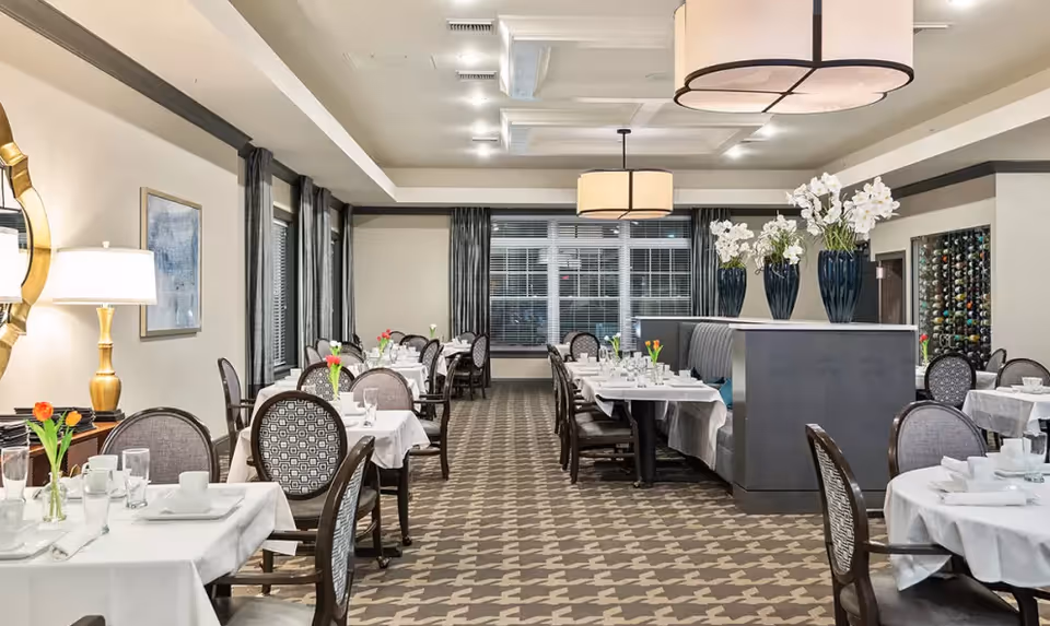 A well-lit dining room with multiple tables covered in white tablecloths, each set with glassware, plates, and napkins. Chairs with patterned upholstery surround the tables. There are large windows with dark curtains, a decorative lamp on a side table, and vases with white flowers on a partition. The carpet has a geometric pattern and a large ceiling light fixture hangs in the center.