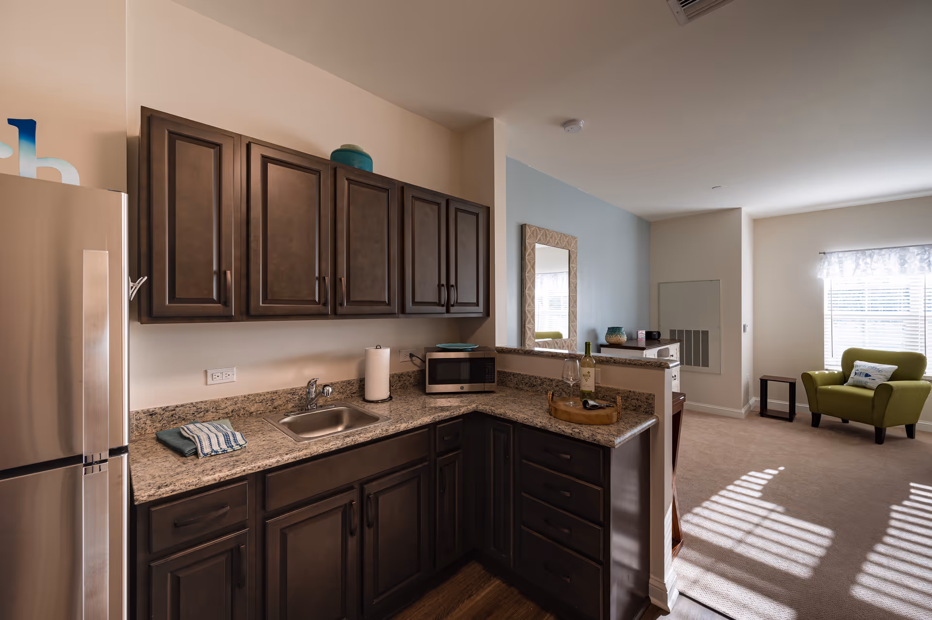 A modern kitchen area with dark wood cabinets, granite countertops, a stainless steel refrigerator, a small sink, a microwave, and a paper towel holder. The kitchen opens into a living space with a green armchair near a window with blinds and a small side table. A decorative mirror hangs on the wall above the counter.