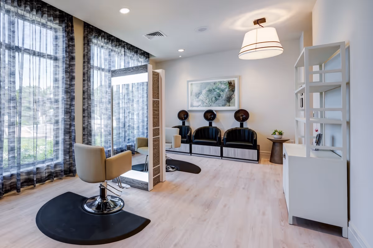 A bright and modern salon area with a styling chair in front of a large mirror, three black hair dryer chairs against the wall, sheer patterned curtains covering large windows, light wood flooring, a white shelving unit, and a contemporary ceiling light fixture.