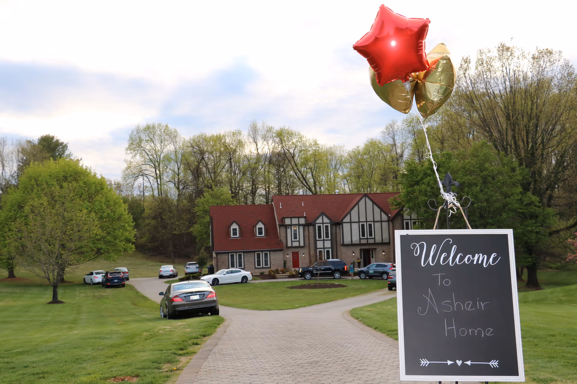 A large house with a red roof and multiple windows is visible at the end of a paved driveway. Several cars are parked on the driveway and lawn. In the foreground, there is a blackboard sign with white cursive writing that says 'Welcome to Asheir Home' and is decorated with red and gold star-shaped balloons. The surrounding area is grassy with trees and a partly cloudy sky.