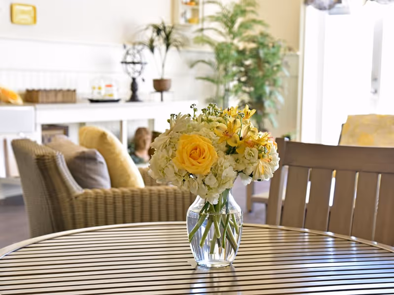 A vase of yellow and white flowers sits on a round wooden table in a bright living room with wicker chairs and plants in the background.