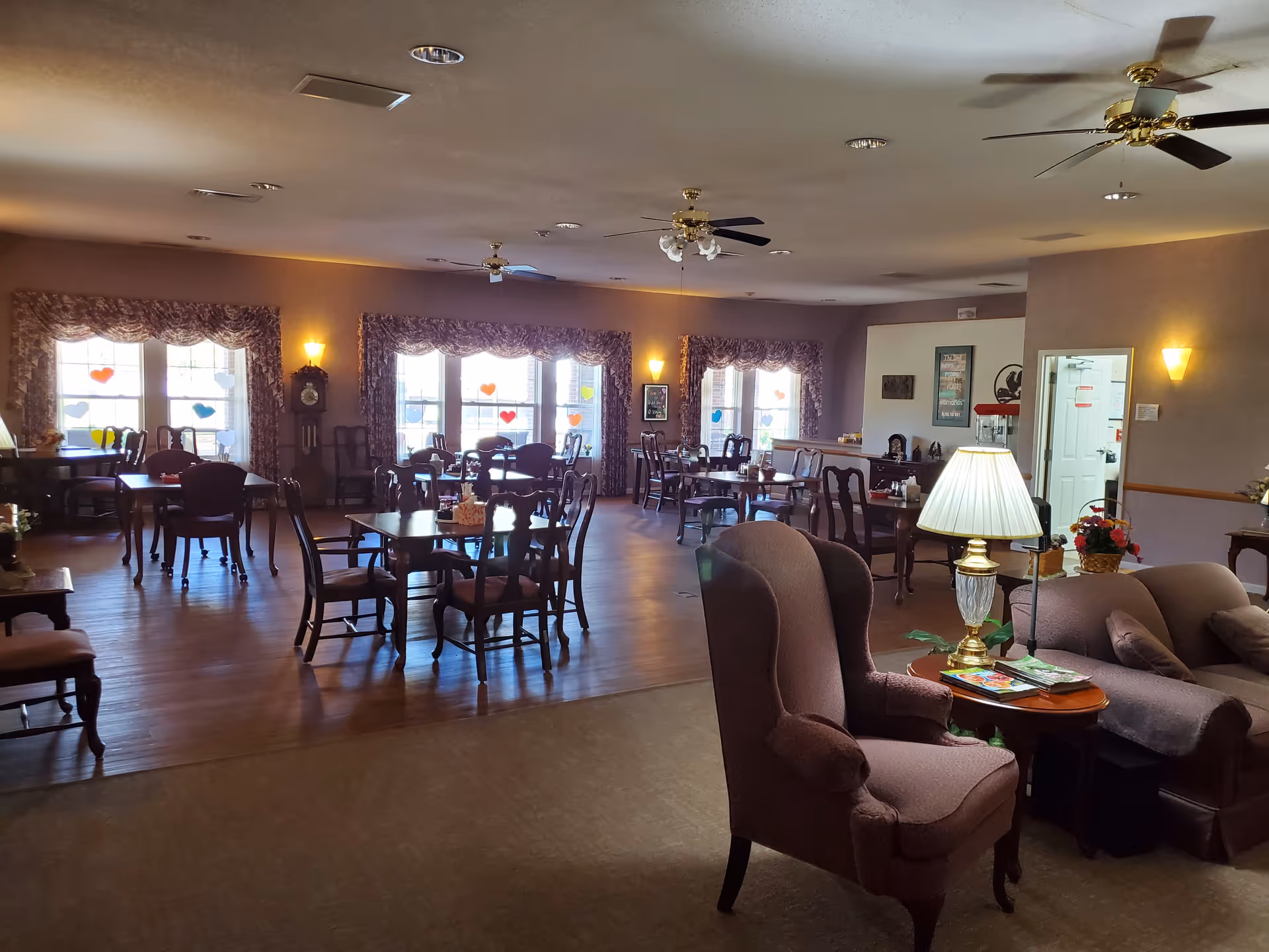 Spacious communal dining room and lounge with multiple tables and chairs, upholstered armchairs, lamps, and large windows with valances.