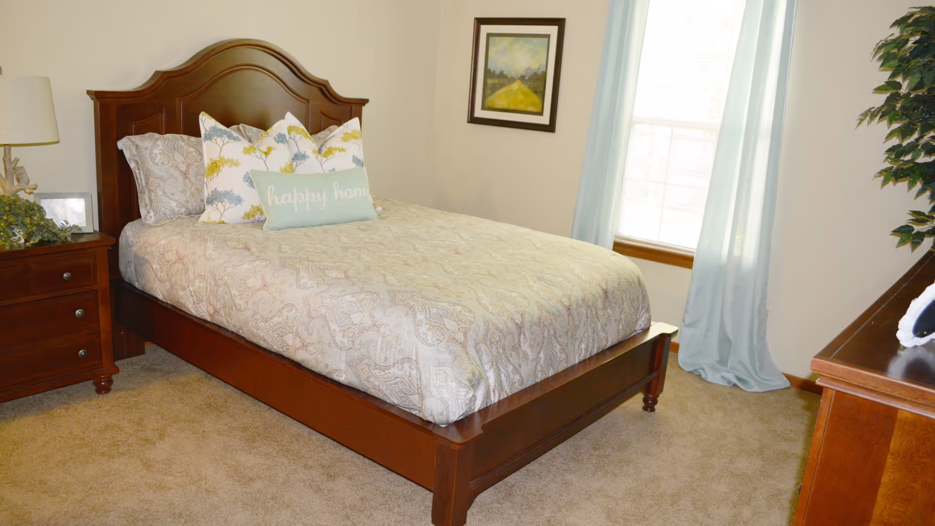 A bedroom with a wooden bed frame and headboard, a bed made with patterned bedding and decorative pillows including one that says 'happy home'. There is a wooden nightstand with a lamp and some greenery on the left side, a framed picture on the wall, a window with light blue curtains, and a wooden dresser on the right side with a plant nearby.