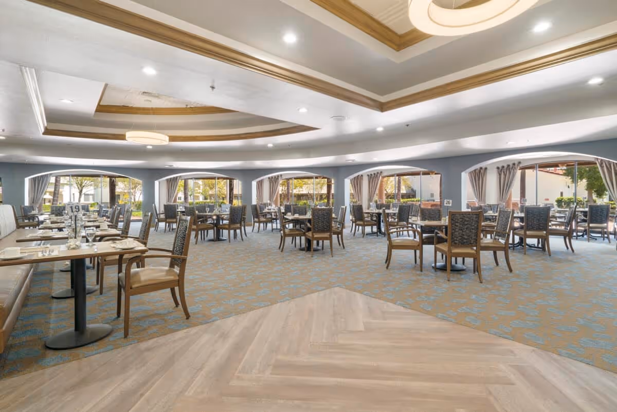 Spacious dining room with multiple tables and chairs arranged neatly. Large windows with curtains allow natural light to fill the room. The ceiling features recessed lighting and decorative wood accents. Tables are set with glasses and napkins, ready for guests.
