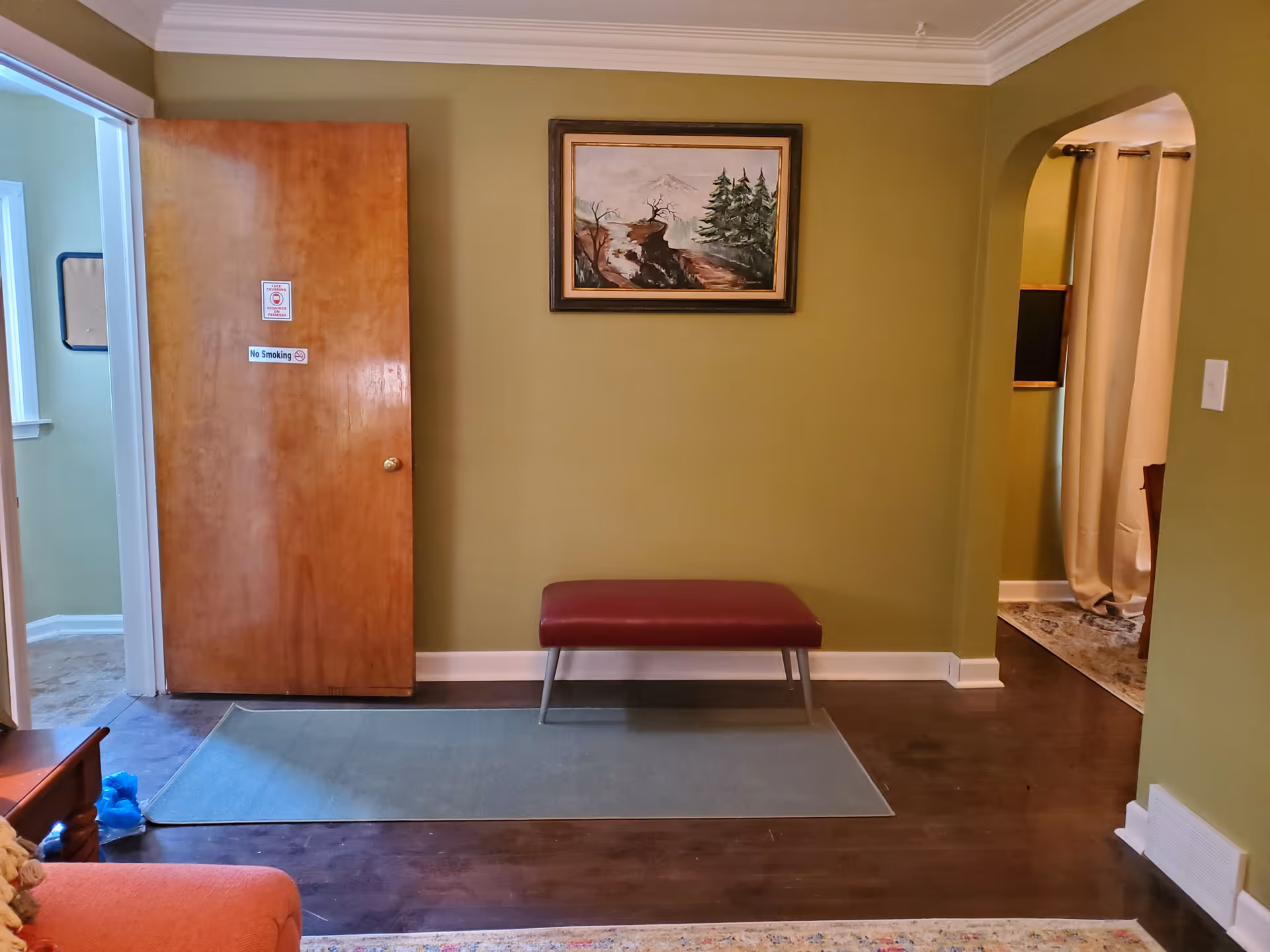 Entryway/living room with olive-green walls, a wooden door on the left, a maroon bench beneath a framed landscape painting and hardwood floors.