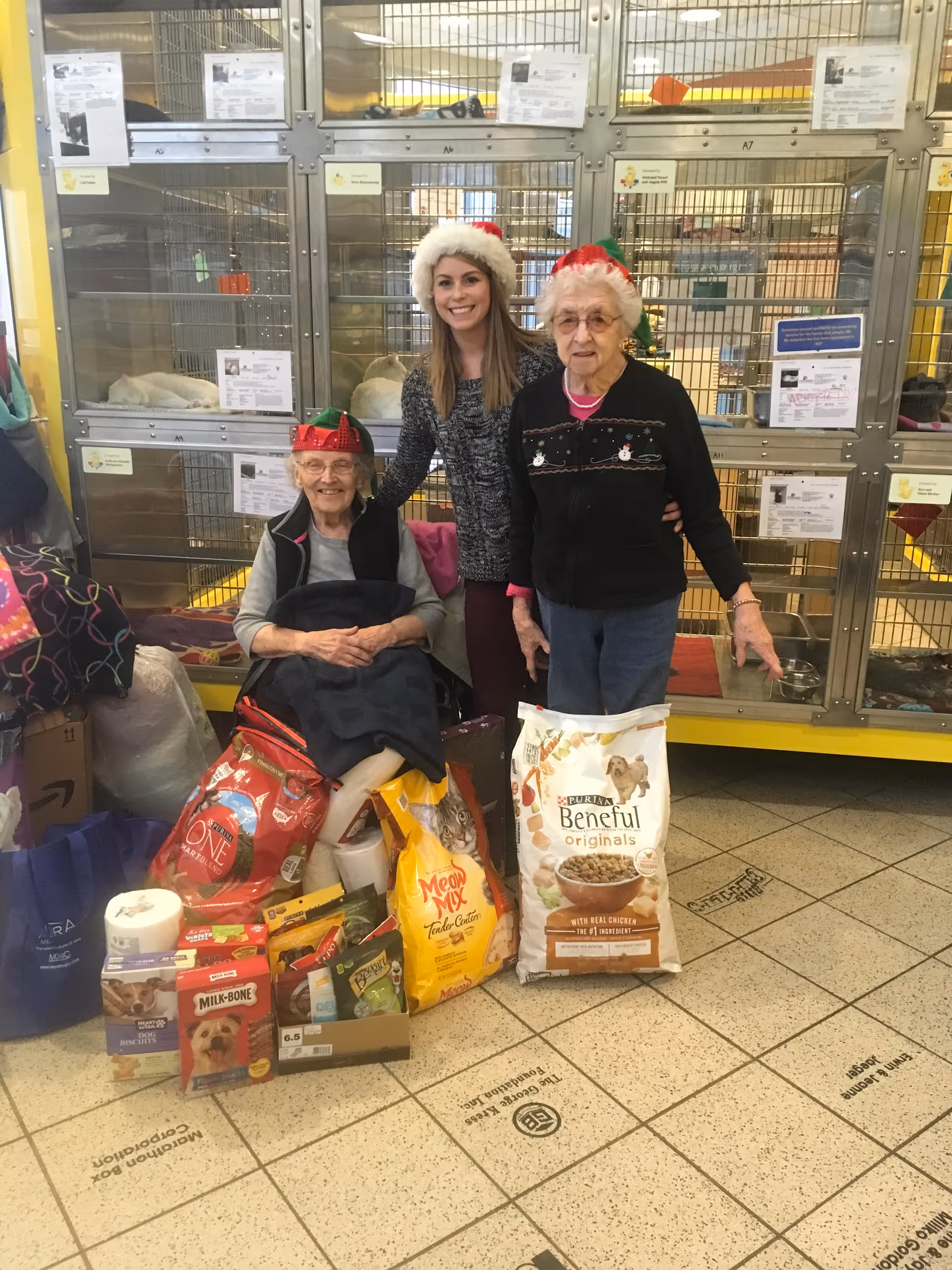 Two elderly women and a younger woman wearing festive holiday hats pose together in front of a row of animal cages. They are surrounded by large bags of pet food and various pet supplies on the floor.