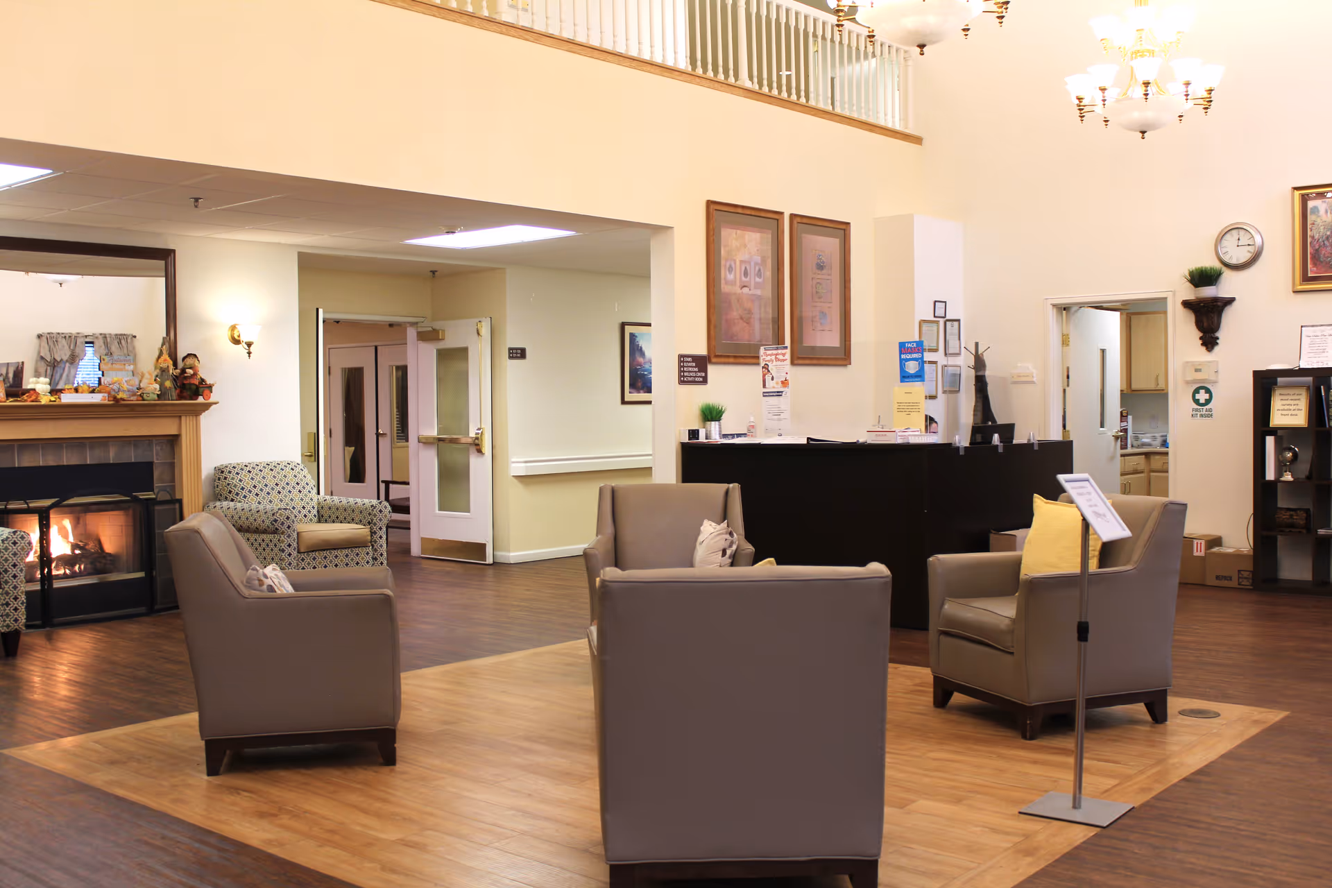 A cozy senior living facility common area with four beige armchairs arranged around a small wooden floor section. There is a fireplace with a lit fire on the left side, decorated with small figurines and a mirror above it. The room has light-colored walls, wooden flooring, and a reception desk in the background with informational posters and a clock on the wall. A chandelier hangs from the ceiling, and there is an open doorway leading to other rooms.