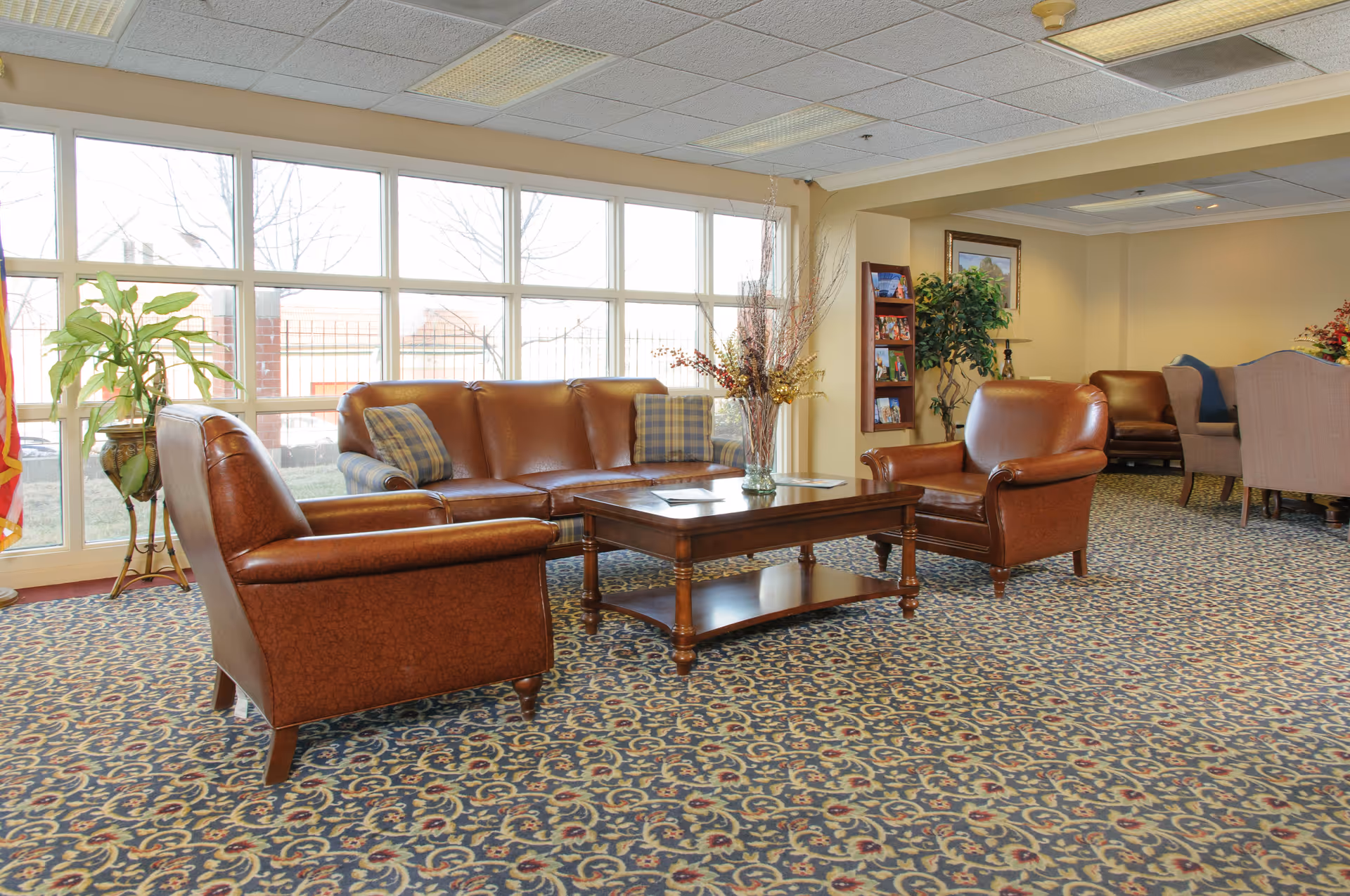 A cozy living room area with large windows letting in natural light. The room features a brown leather sofa and two matching armchairs arranged around a wooden coffee table with a floral centerpiece. The carpet has a detailed patterned design, and there are plants and framed pictures decorating the space.
