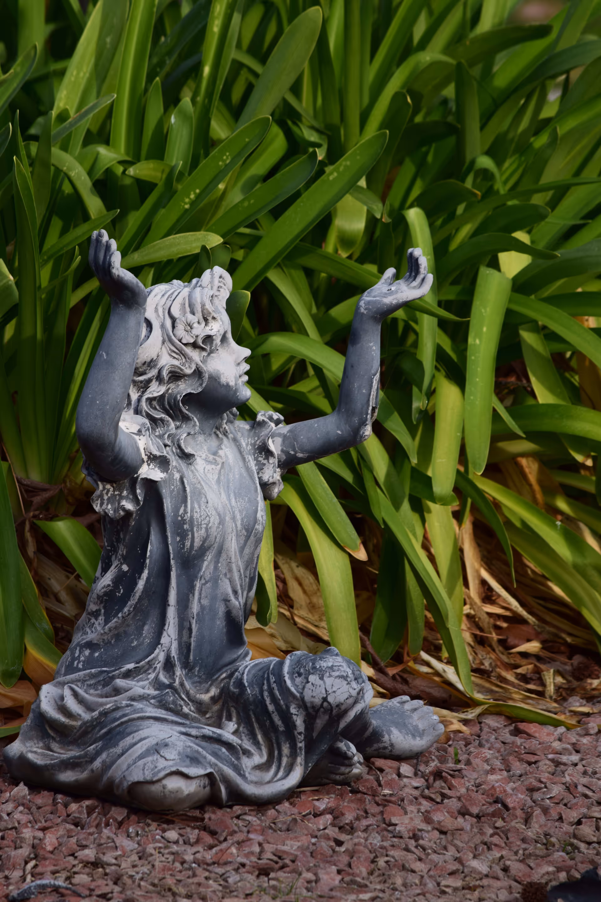 A weathered gray statue of a young girl sitting cross-legged on reddish gravel with her arms raised, surrounded by lush green leafy plants in the background.