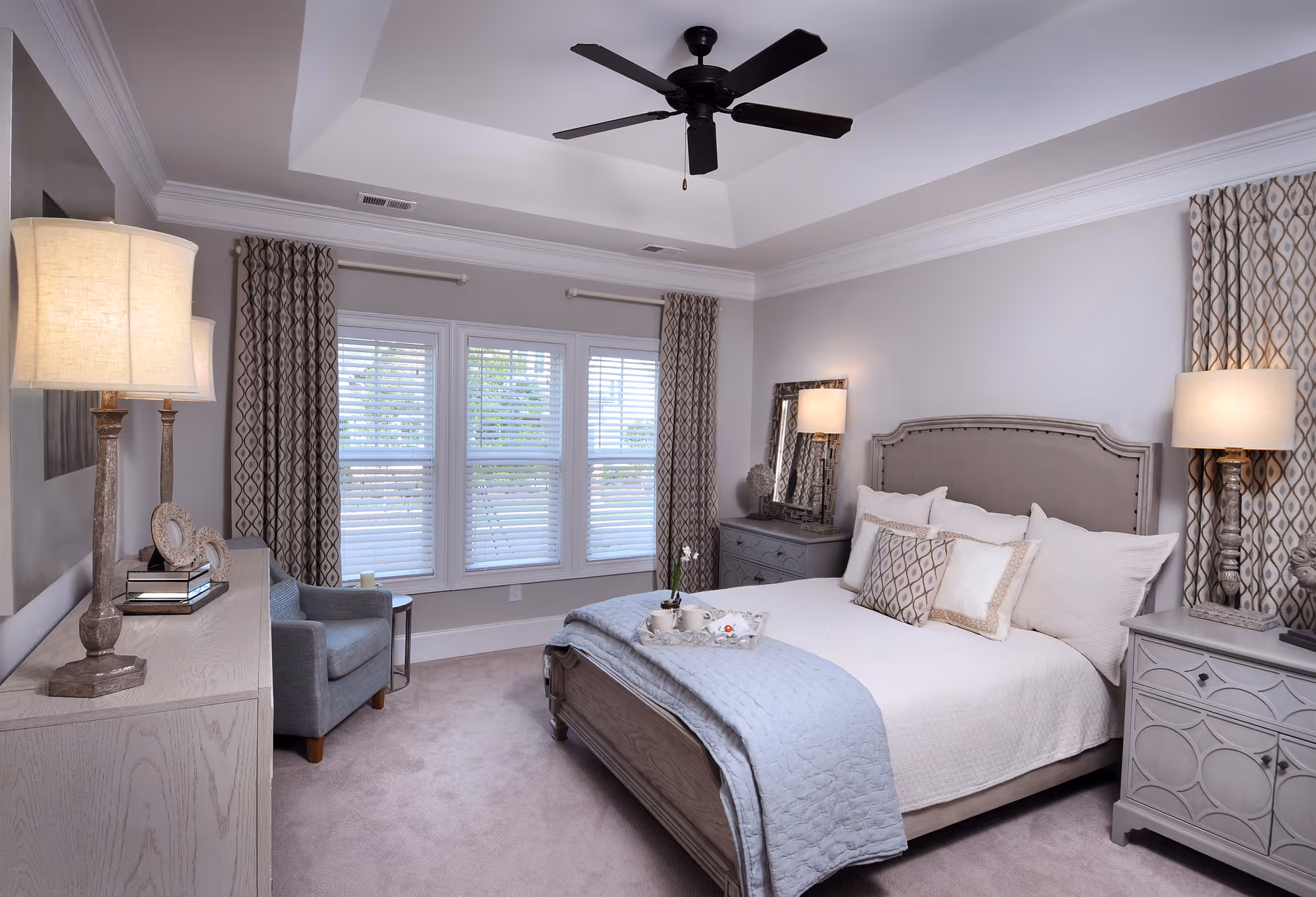 A well-lit bedroom with a large bed featuring multiple pillows and a light blue quilt. The room has beige walls, a ceiling fan, patterned curtains on windows with white blinds, two bedside tables with lamps, a dresser with decorative items, and a comfortable armchair in the corner.