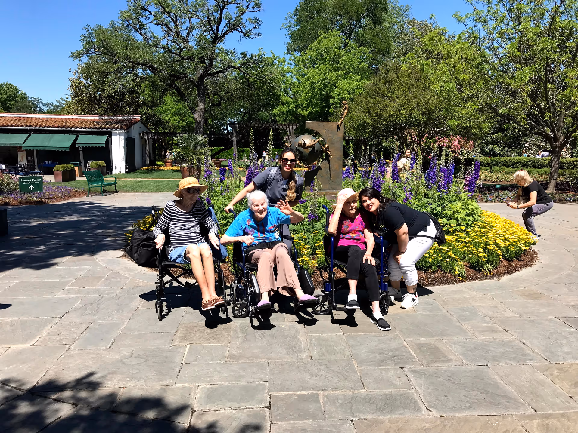 A group of elderly women, some in wheelchairs, and two caregivers are posing and smiling in front of a flower bed with purple and yellow flowers in an outdoor garden area on a sunny day. There is a bronze sculpture behind them and trees and a building with a green awning in the background.