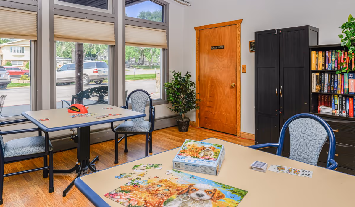 Bright activity/common area in a senior living facility with tables, chairs, a bookshelf, and large windows.