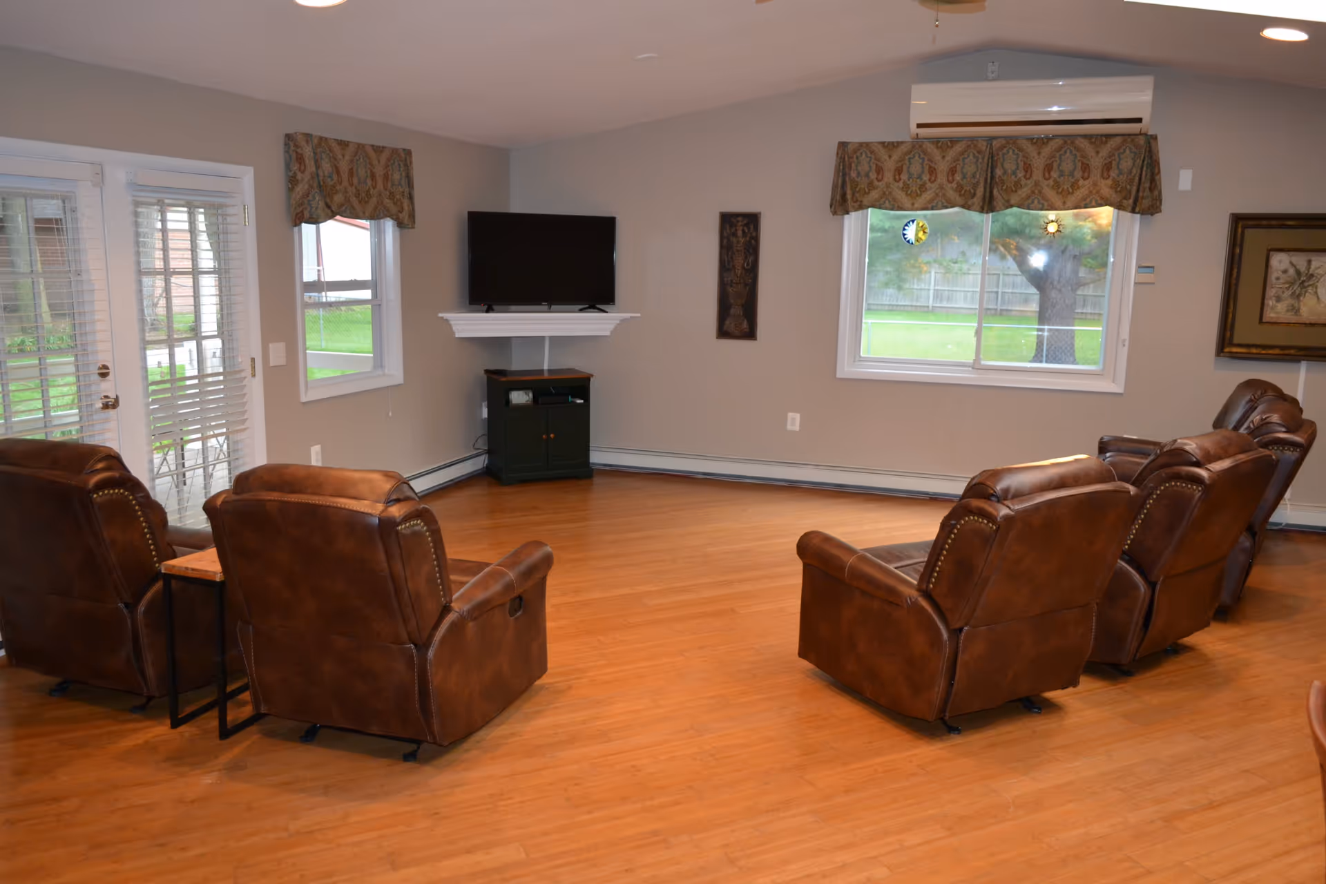 A living room with four brown leather recliners arranged in two rows facing a wall-mounted flat screen TV. The room has wooden flooring, two windows with patterned valances, a glass door with blinds, and a small black cabinet under the TV. There is an air conditioning unit above one window and framed artwork on the walls.