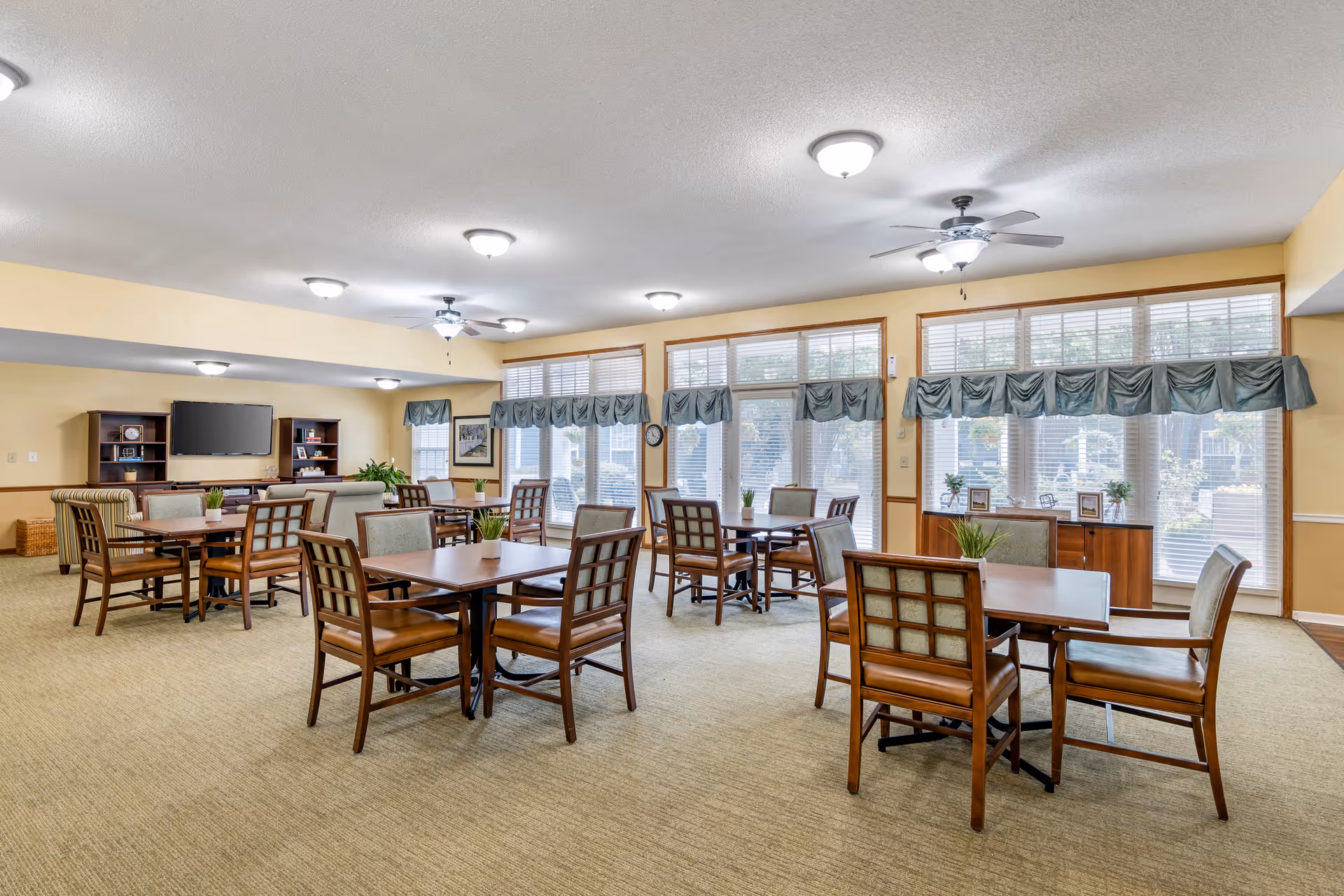 A spacious, well-lit common area with multiple wooden tables and chairs arranged neatly. Large windows with blue valances allow natural light to fill the room. The walls are painted a soft yellow, and there is a TV mounted on a wooden entertainment unit in the background. The room has ceiling fans and overhead lights, creating a comfortable and inviting atmosphere.