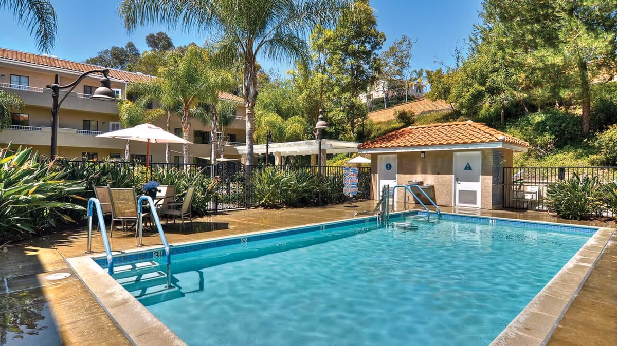 Outdoor swimming pool area at a senior living facility with clear blue water, metal handrails, surrounding patio with tables, chairs, umbrellas, palm trees, and a small building with restroom doors in the background.