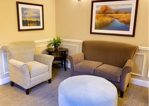 A cozy sitting area with a beige armchair, a brown loveseat, a round light blue ottoman, and a small dark wooden side table with a green plant. Two framed landscape paintings hang on the cream-colored walls above the seating.