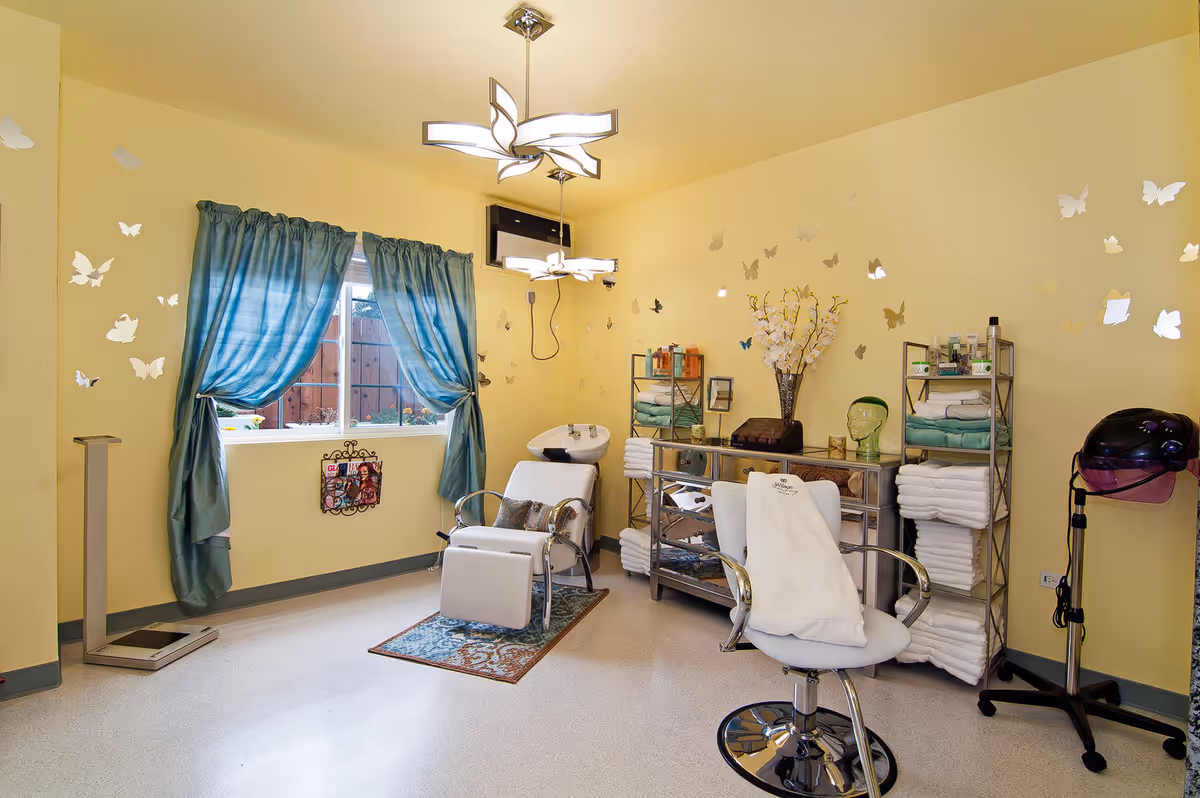 A bright room with yellow walls decorated with butterfly decals, featuring two salon chairs, one with a towel draped over it, a hair washing station, shelves stocked with towels and hair care products, a window with blue curtains, and a hair dryer on a stand.