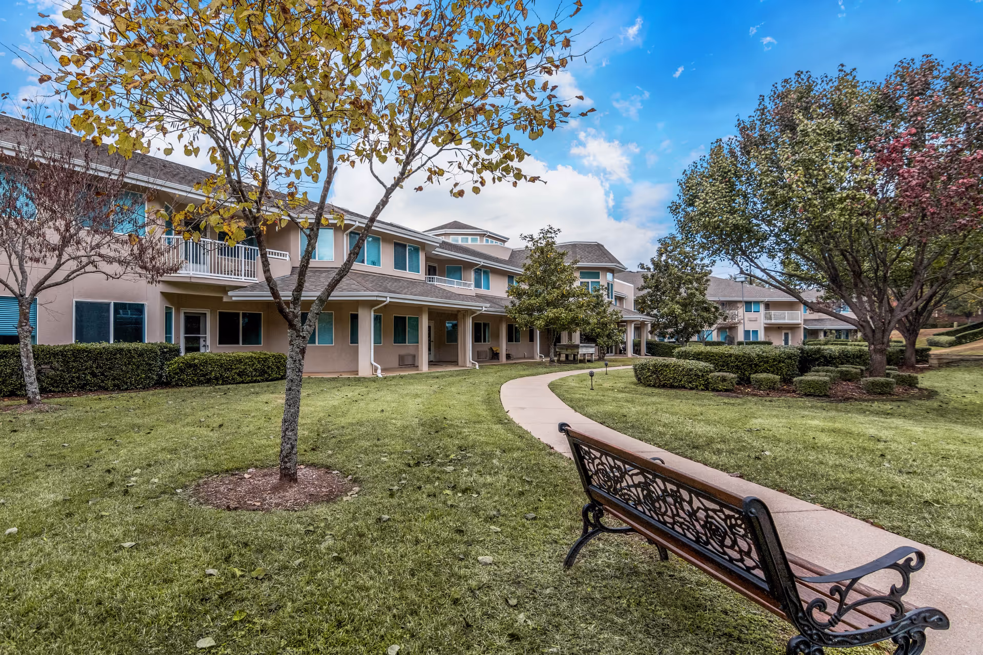 Outdoor view of Asher Point Independent Living of Tyler showing a well-maintained grassy area with a curved concrete pathway, a decorative metal bench in the foreground, several trees with autumn leaves, and a two-story building with multiple windows and balconies in the background under a partly cloudy blue sky.
