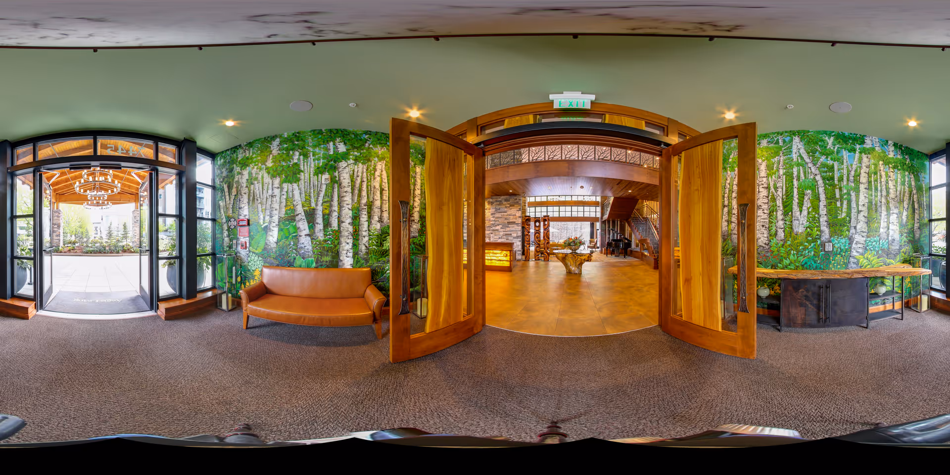 Entrance area of Aegis Living Mercer Island featuring large wooden double doors opening into a spacious interior with a wooden table and decorative elements. The walls on either side of the entrance are adorned with a mural of birch trees and greenery. There is a brown leather bench on the left side and a console table on the right side near large windows. The entrance leads to a well-lit interior space with wooden and stone accents.