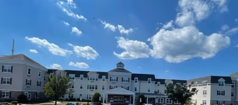 Exterior view of a large, multi-story senior living facility building under a blue sky with scattered clouds. The building has a central entrance with a covered driveway and multiple windows, surrounded by some trees and a parking area.