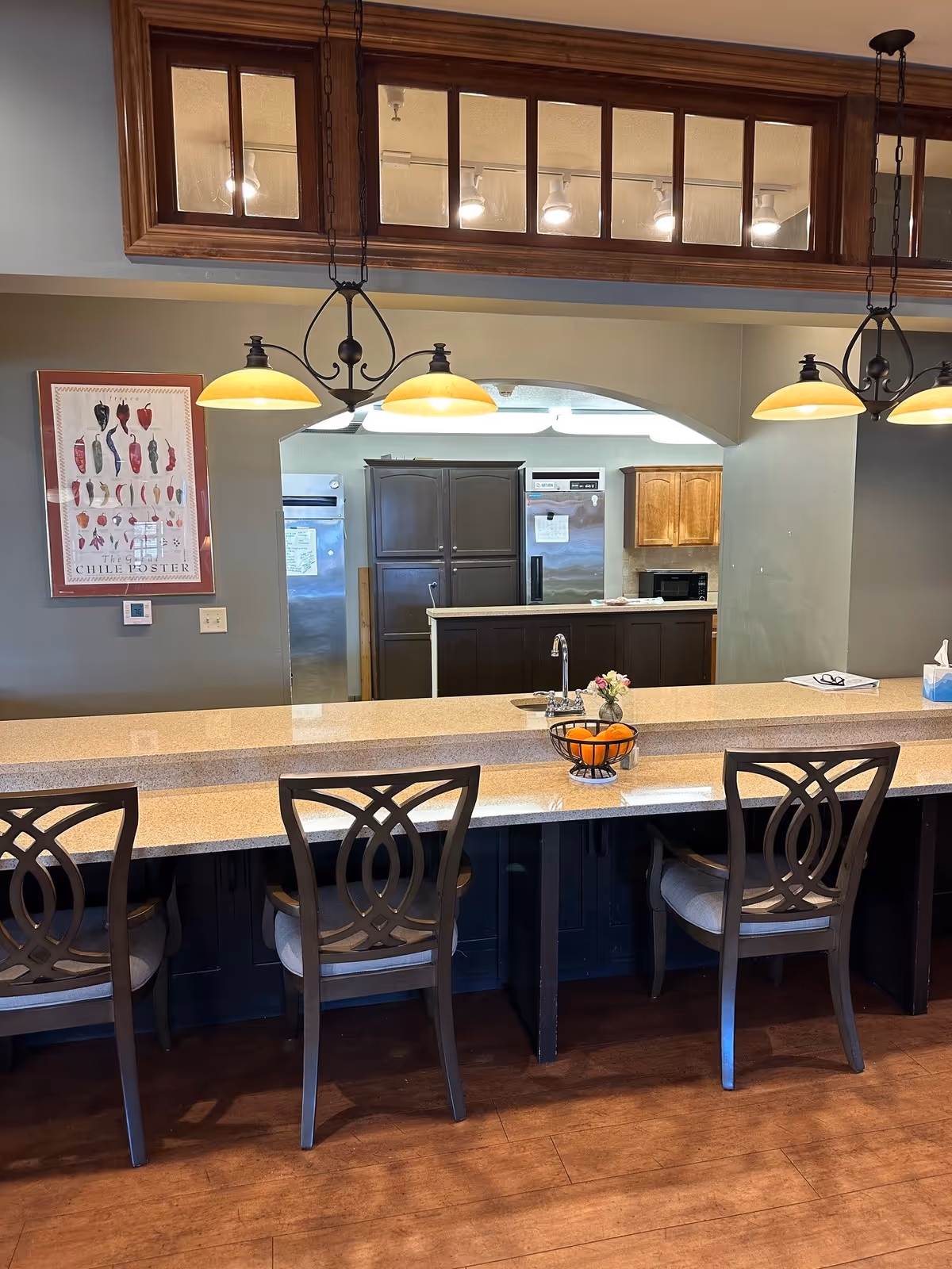 Interior view of a kitchen area with a long counter and four chairs with decorative backs. Above the counter are three hanging pendant lights with yellow shades. Behind the counter, there is a kitchen with dark and light wooden cabinets, a stainless steel refrigerator, and a microwave. A framed poster of chili peppers is on the wall to the left.