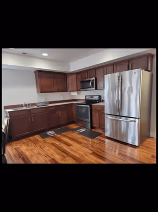 Modern kitchen with wooden cabinets, stainless steel refrigerator, oven, microwave, and a double sink. The floor is polished wood and there are two black floor mats in front of the sink and stove.