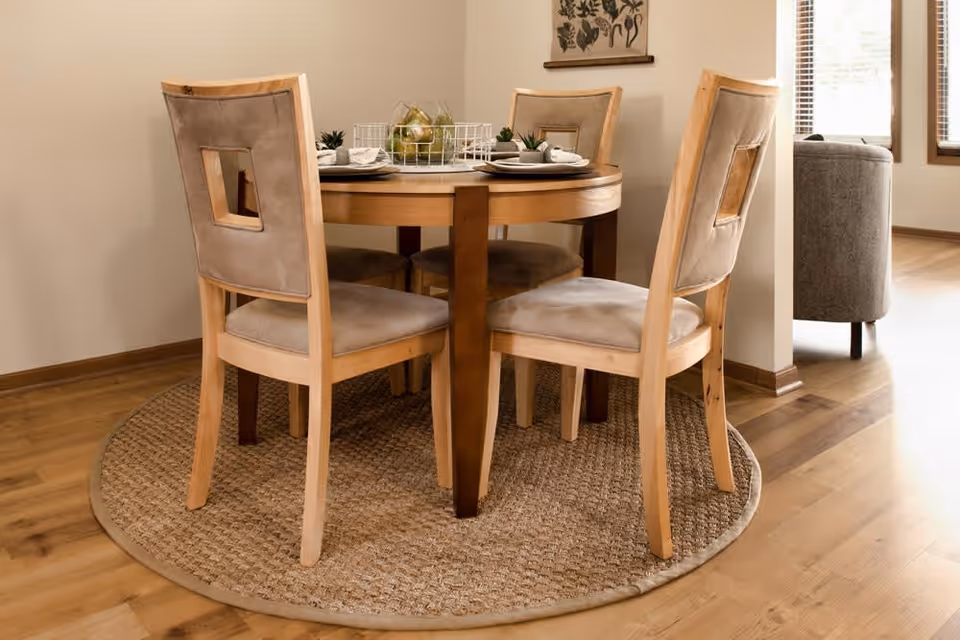 A round wooden dining table with four cushioned chairs arranged around it, placed on a circular woven rug. The table is set with plates, napkins, and small decorative plants. The room has wooden flooring and neutral-colored walls, with a window and a gray armchair visible in the background.