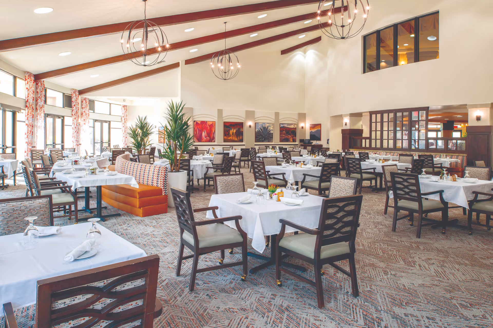 A spacious and well-lit dining room with multiple tables covered in white tablecloths, set with napkins and glassware. The room features high vaulted ceilings with modern spherical chandeliers, large windows with patterned curtains allowing natural light, and decorative plants. There are various seating options including chairs and cushioned benches, with colorful artwork on the walls.