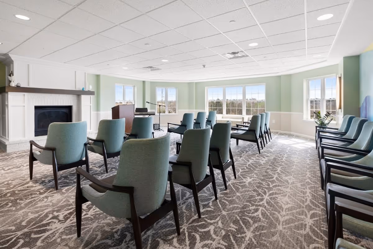 A bright meeting or activity room with multiple rows of light blue upholstered chairs arranged facing a podium and microphone stand. The room has large windows letting in natural light, a white fireplace on the left, and patterned carpet flooring.
