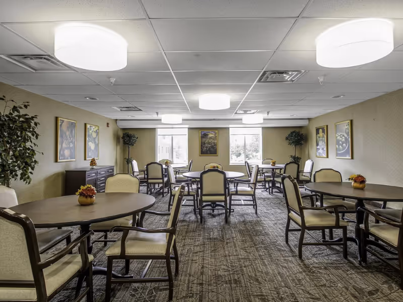 A well-lit dining room with several round tables and cushioned chairs arranged neatly. The room has carpeted flooring, beige walls adorned with framed artwork, and large windows letting in natural light. There are also potted plants placed in the corners of the room.
