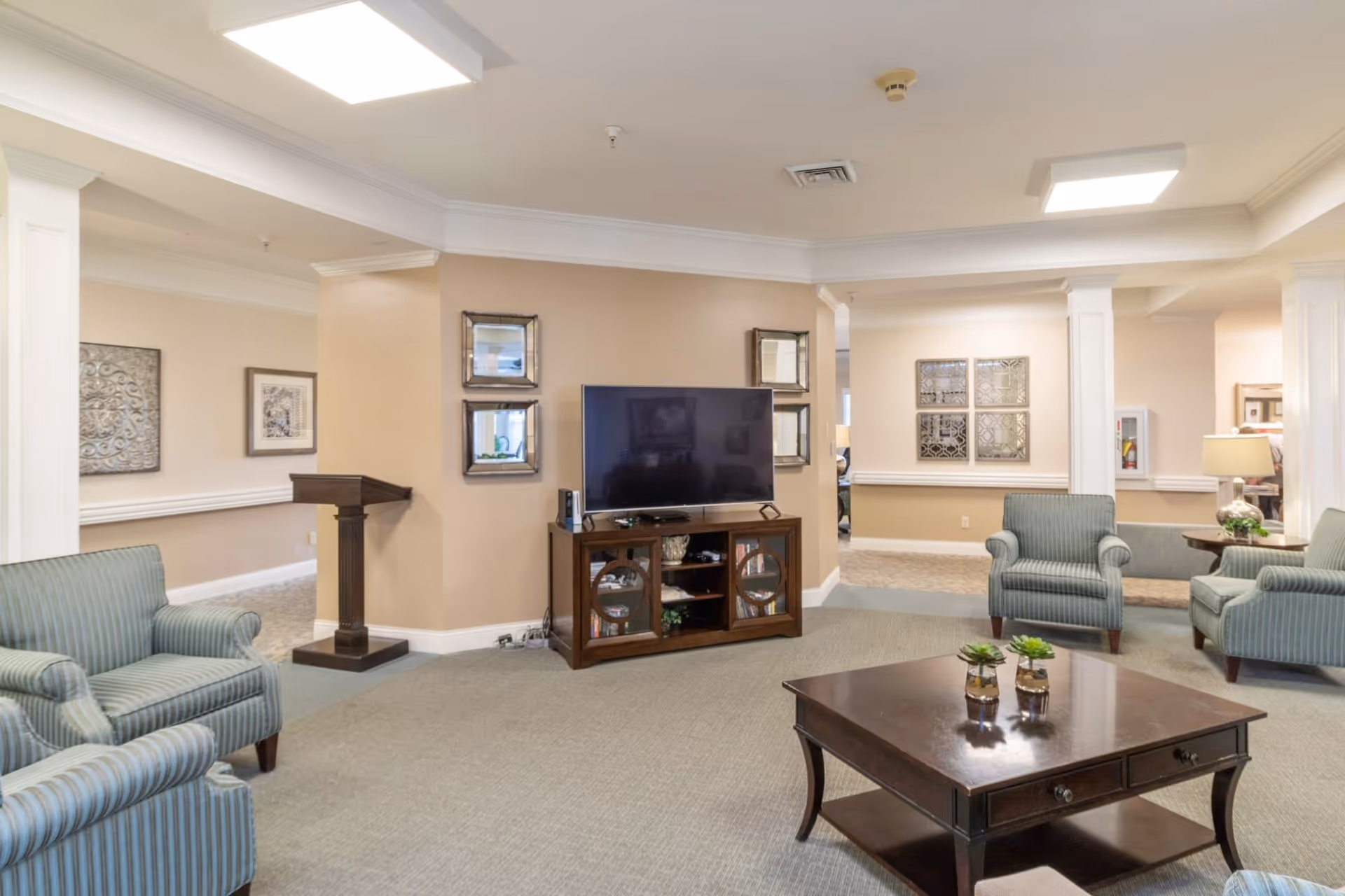 A cozy living room area in a senior living facility featuring a flat-screen TV on a wooden cabinet, four striped armchairs arranged around a dark wooden coffee table with small potted plants, beige walls with decorative mirrors and artwork, and soft overhead lighting.