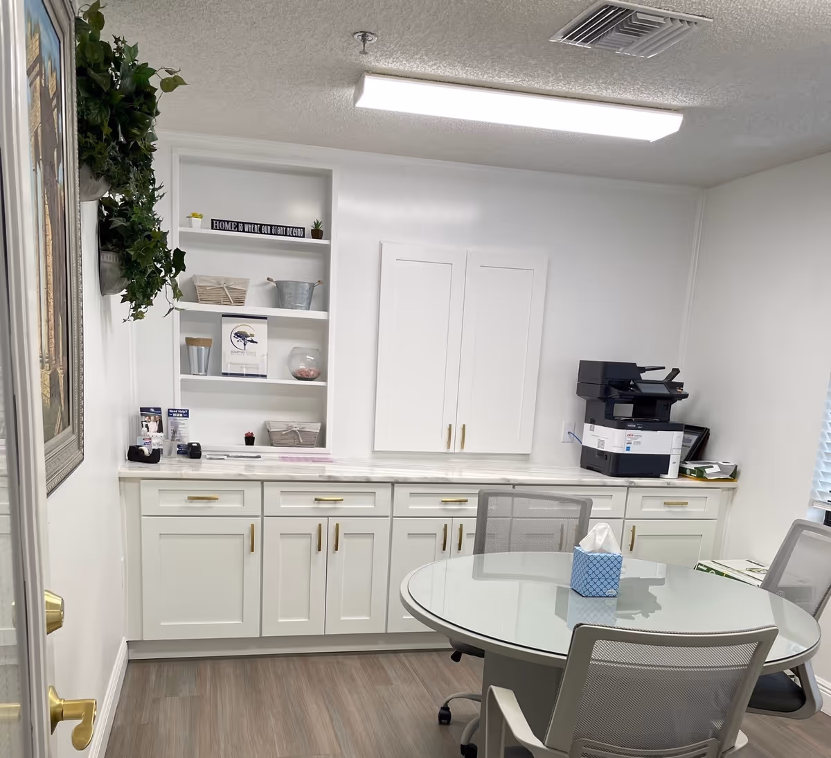 A small office or meeting room with white cabinets and shelves against the wall, a round glass table with mesh office chairs around it, a tissue box on the table, a printer on the countertop, and some decorative plants and items on the shelves.