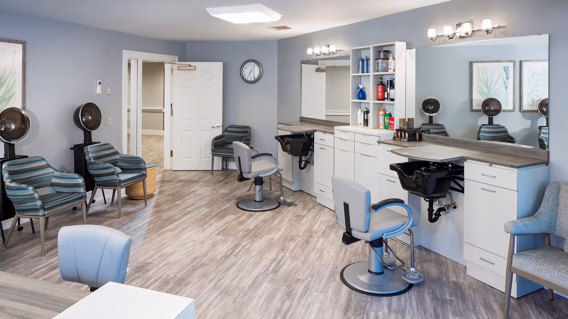 A clean and modern salon area in a senior living facility with multiple styling chairs, hair washing stations, and hair dryers. The room has light gray walls, wood-look flooring, a large mirror above the counter with various hair products, and comfortable seating for waiting.