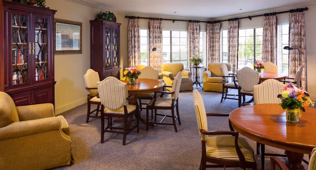 A cozy and well-lit common area with round wooden tables surrounded by striped upholstered chairs. There are comfortable armchairs near large windows with floral curtains, and flower arrangements on the tables. A wooden cabinet with glass doors and books is visible on the left wall.