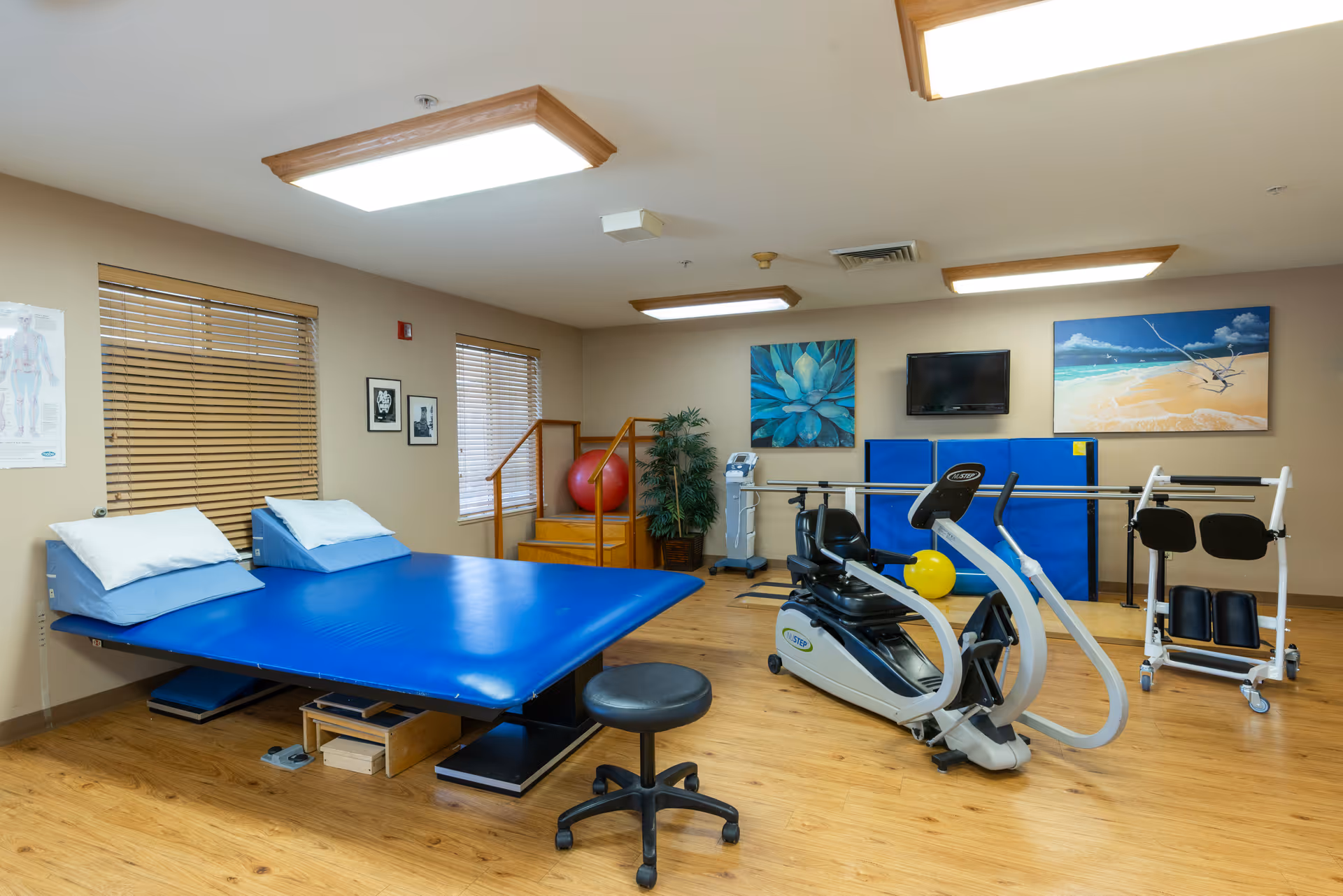 A bright physical therapy room with a blue treatment table, exercise bike, therapy equipment, and coastal wall art.