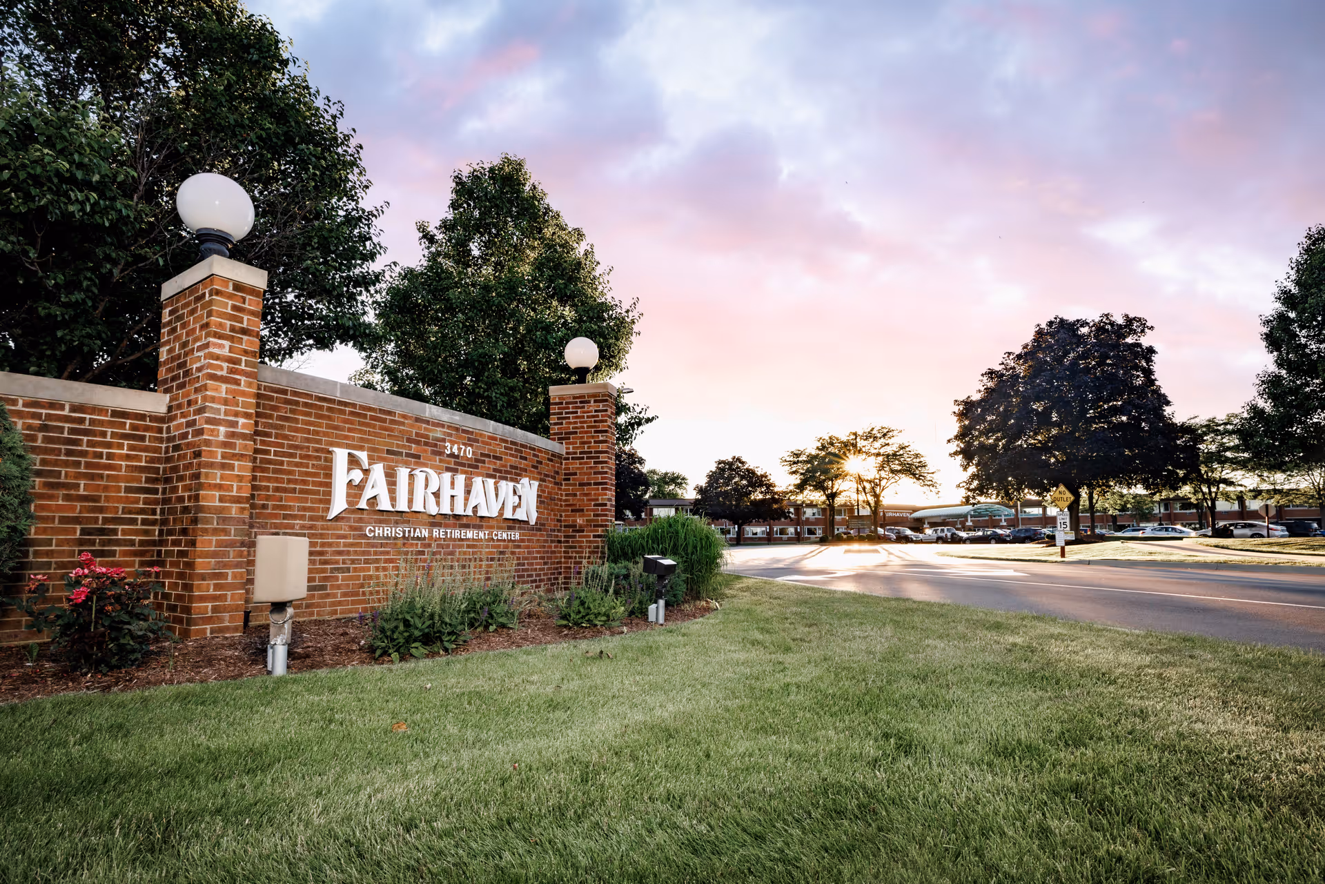 Entrance sign for Fairhaven Christian Retirement Center made of brick with white lettering, surrounded by green grass, plants, and trees, with a road and parking lot visible in the background under a partly cloudy sky at sunset.