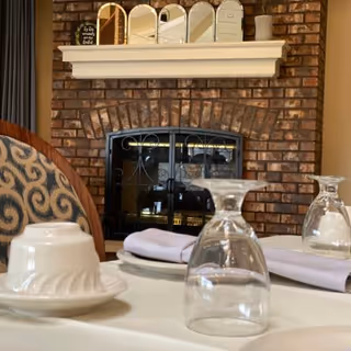 A close-up view of a dining table set with upside-down wine glasses, white plates, and folded napkins in front of a brick fireplace with a white mantel. Decorative items including small mirrors and containers are placed on the mantel.