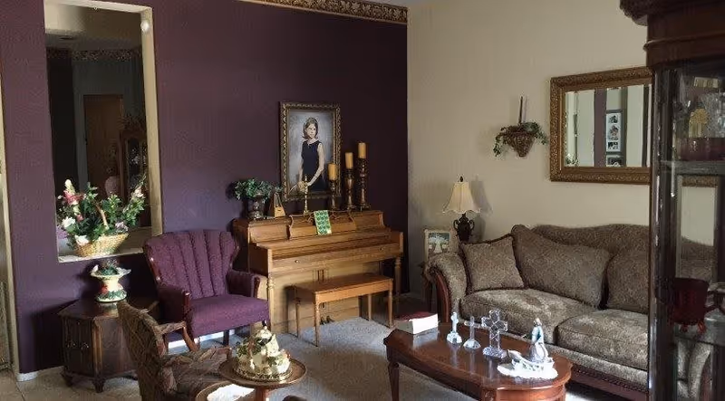 A cozy living room with a brown patterned sofa, a wooden coffee table with decorative items, a wooden piano against a purple accent wall with a framed portrait above it, a purple upholstered armchair, a side table with a basket of flowers, and a large mirror on the adjacent beige wall.