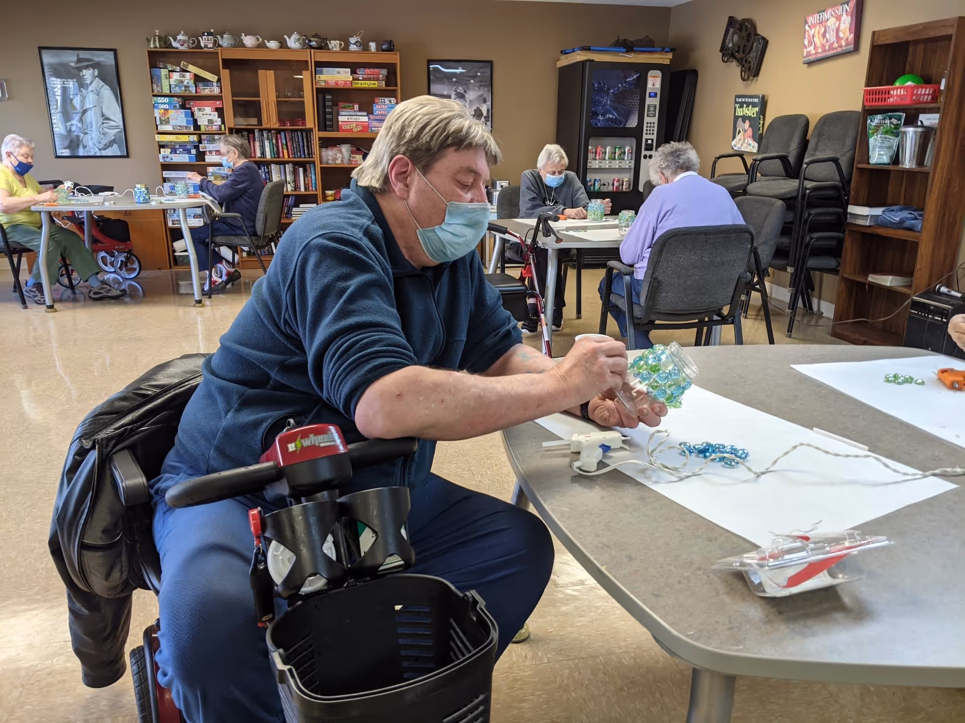 Several elderly individuals wearing face masks are seated at tables in a senior living facility activity room, engaging in arts and crafts. One person in the foreground is decorating a jar with colorful glass beads. The room has shelves filled with board games and books, a vending machine, and posters on the walls.