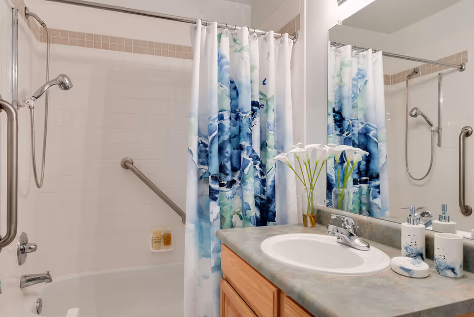 Bathroom with a bathtub and shower combination featuring a silver grab bar and a shower curtain with blue and green floral patterns. A countertop with a sink, faucet, and decorative items including a vase with white flowers and matching soap dispensers is visible. A large mirror reflects part of the bathroom.