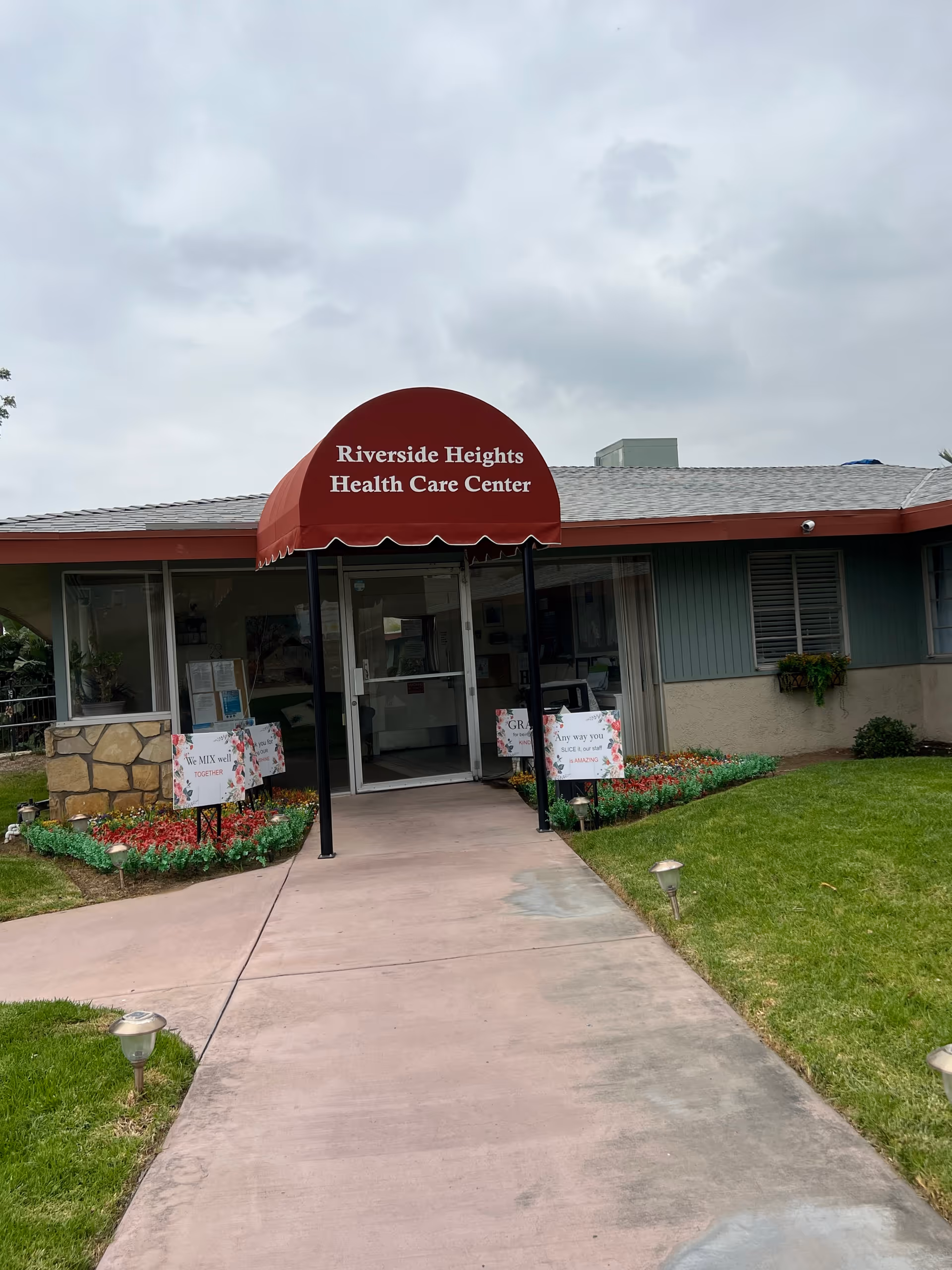 Entrance to Riverside Heights Health Care Center with a red awning displaying the facility name. The pathway leading to the door is flanked by green grass and flower beds with small signs. The building has a light blue exterior with windows and a stone accent wall on the left side.