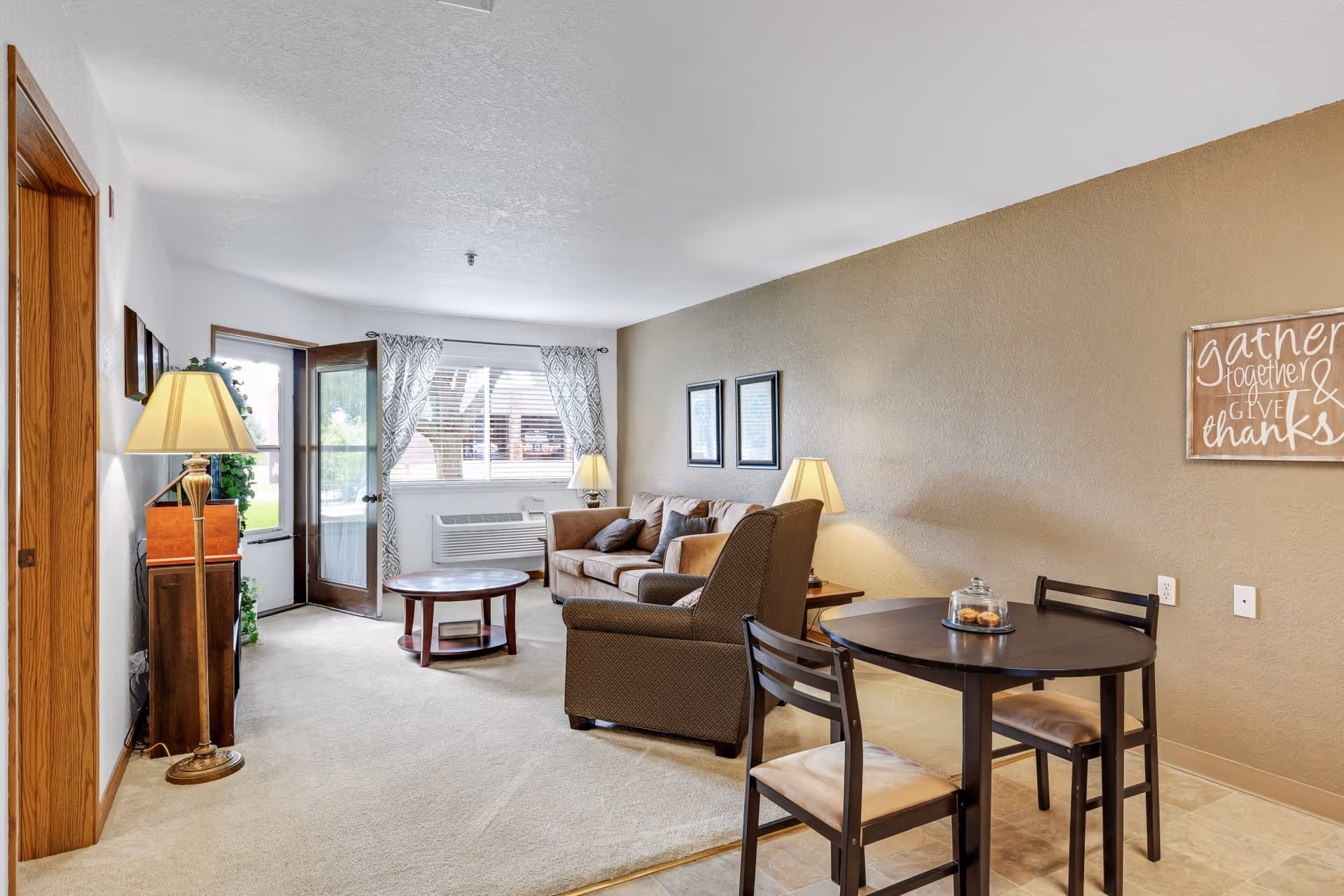 Cozy living room and small dining area with a sofa, armchair, coffee table, round dining table, lamps, window and an exterior door.