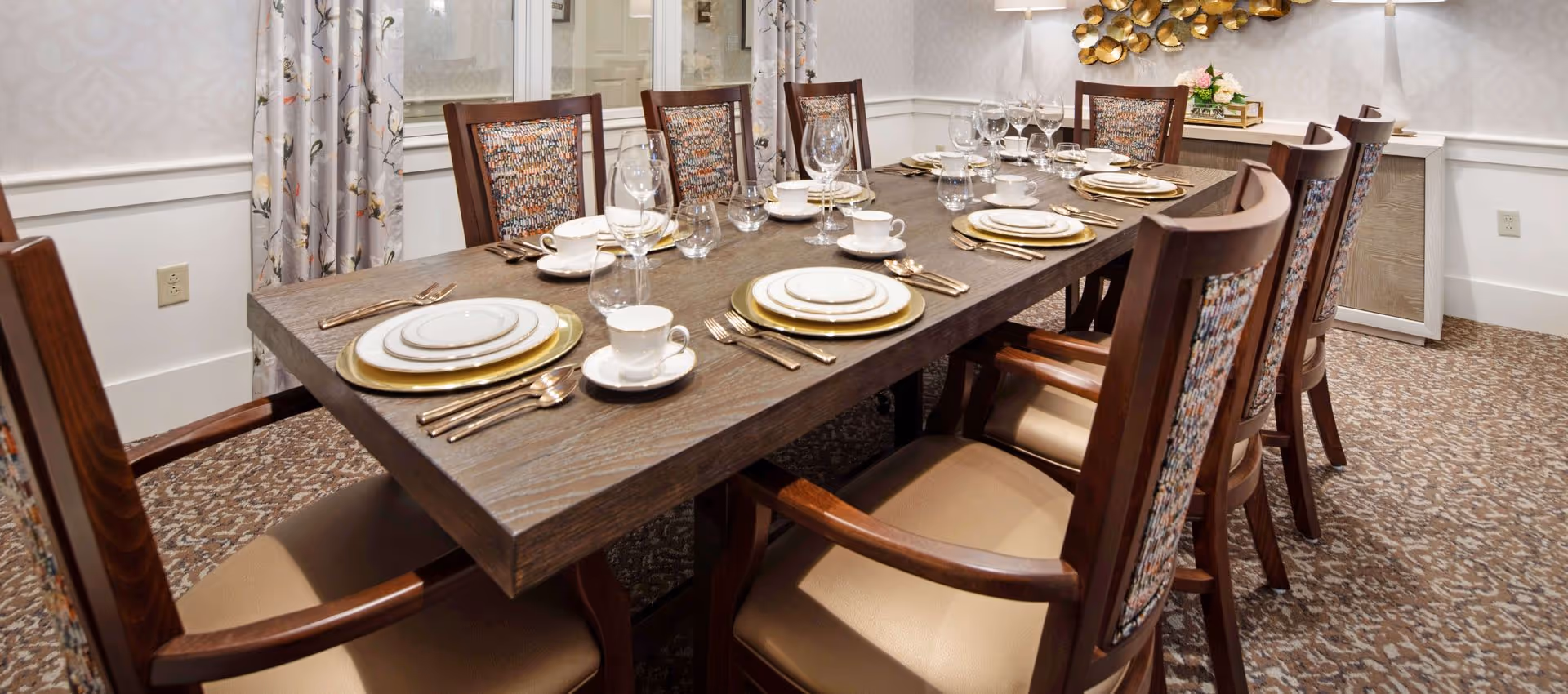 A formal dining table set with plates, glassware, cutlery and upholstered chairs in a decorated dining room.